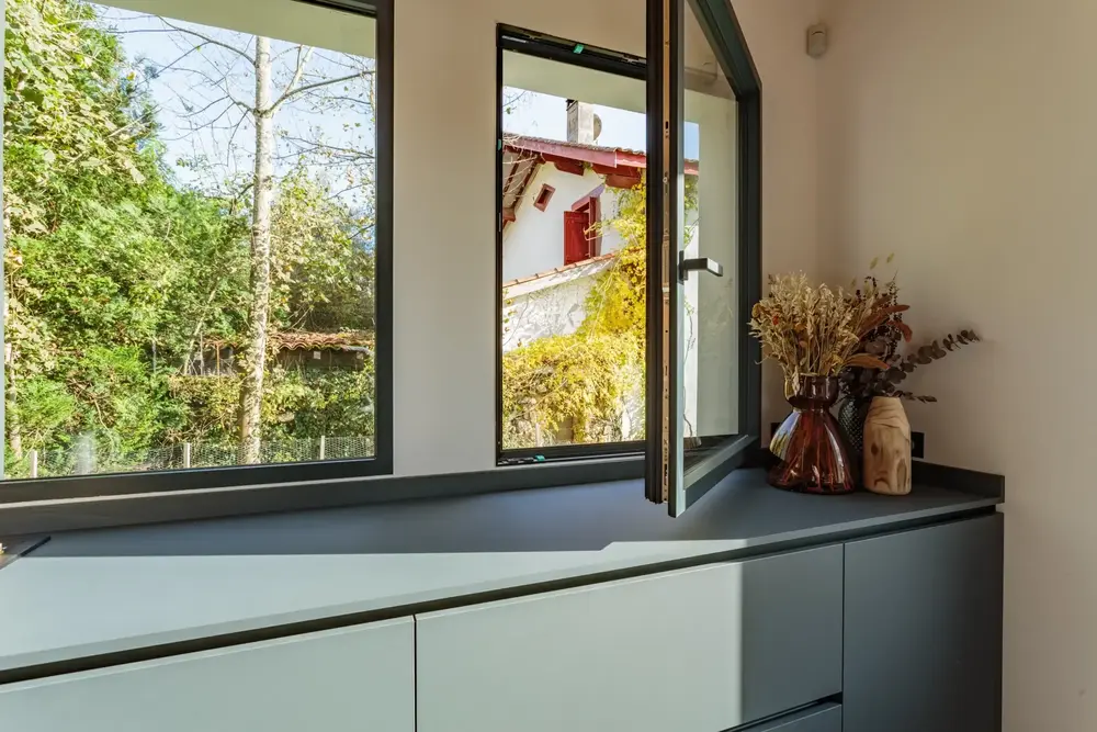 Fenêtre aluminium oscillo-battante noire ouverte en vue intérieure, avec un aperçu d'une maison traditionnelle de la région de Pau