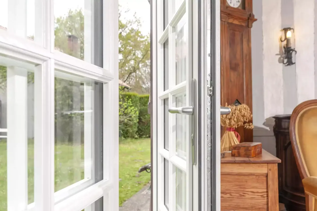 Gros plan du mécanisme et de la poignée en inox d'une porte-fenêtre en bois blanc à petits carreaux, s'ouvrant sur le jardin