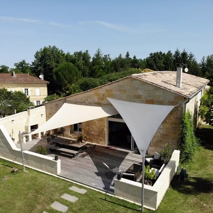 Vue aérienne d'une terrasse de maison ancienne béarnaise avec deux voiles d'ombrage triangulaires blancs fixées sur des murs en pierre.