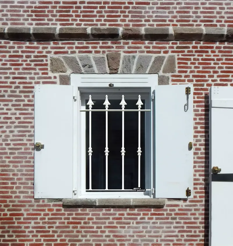 Grille de défense en fer forgé modèle Victoria, couleur blanche, installée sur fenêtre de maison ancienne en briques rouges.