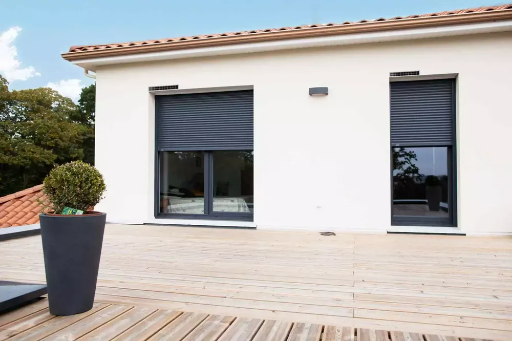 Installation de volets roulants solaires gris sur une façade blanche, créant un contraste moderne sur une terrasse en bois.
