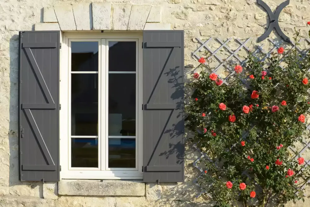 Volets battants gris sur un mur en pierre, façade traditionnelle de maison béarnaise et roses grimpantes.